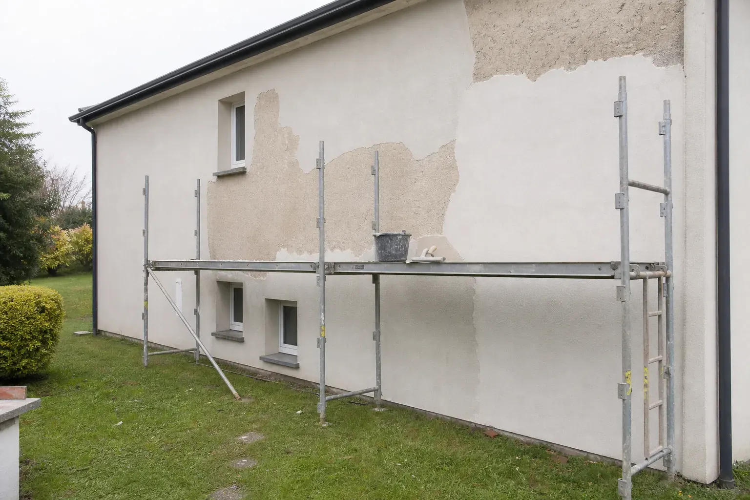 Façade de maison en cours de remise en état, plages d’enduit dégarnies et réparation plus claire, échafaudage de pied le long du mur