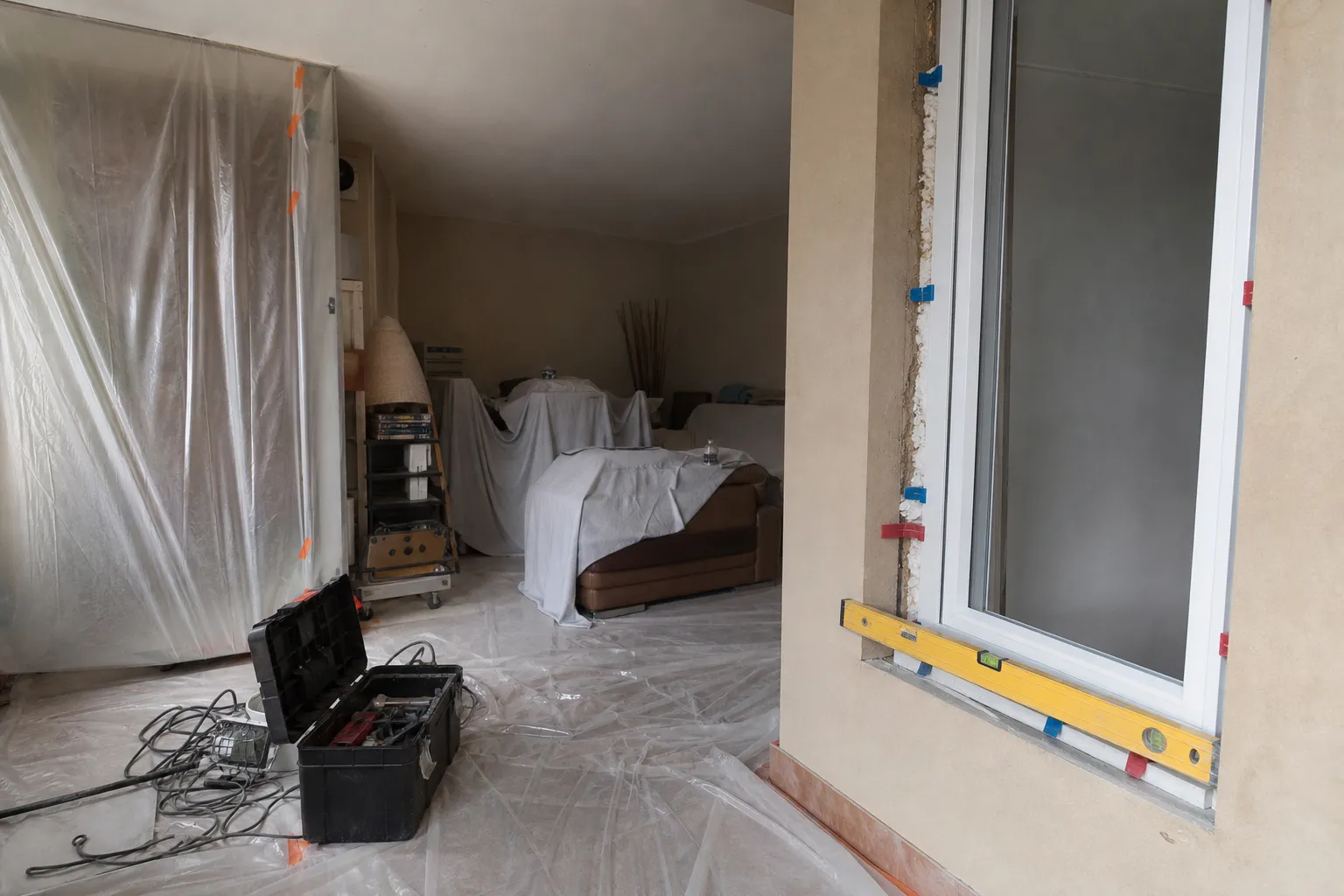 Pose d’un dormant de fenêtre dans un séjour habité, bâche de séparation tendue et meubles regroupés sous des draps de protection