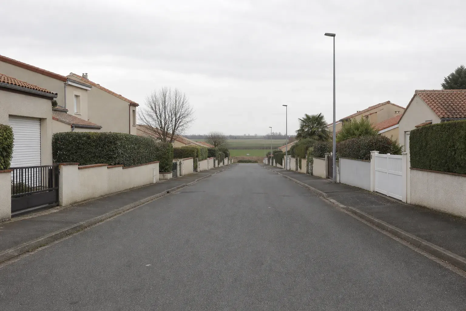 Rue de lotissement pavillonnaire enduit des années 1990-2010, portails et haies basses, plaine cultivée au bout de la rue