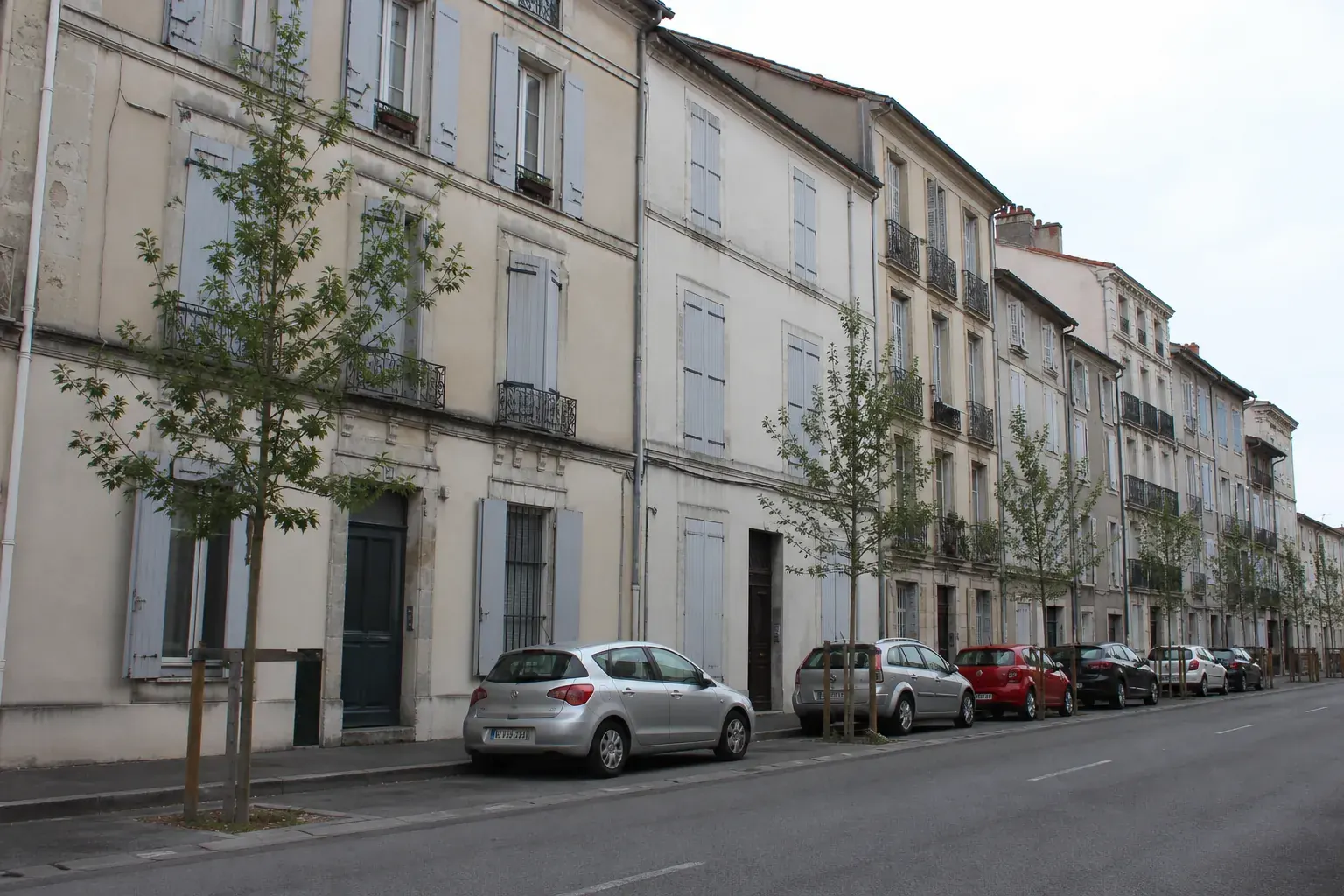 Rue de ville-centre bordée d’immeubles d’habitation mitoyens à volets battants, sous un ciel blanc laiteux